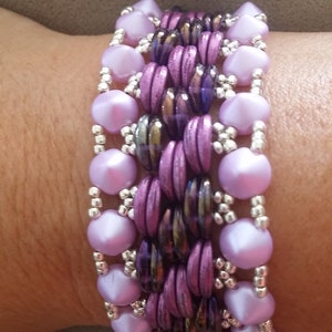 Puede incluir: Un brazalete de múltiples hilos con cuentas moradas y plateadas. El brazalete está hecho con una variedad de cuentas, incluyendo cuentas pequeñas redondas, cuentas facetadas más grandes y cuentas pequeñas plateadas.