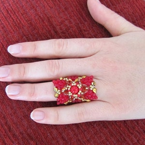 May include: A red and gold beaded ring with a large red gemstone in the center. The ring is worn on a finger.