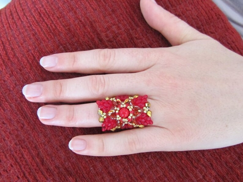 May include: A red and gold beaded ring with a large red gemstone in the center. The ring is worn on a finger.