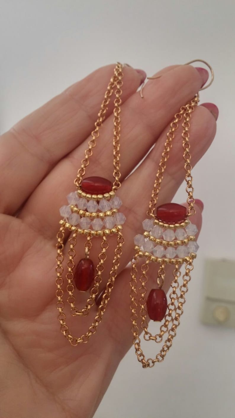 May include: A pair of long gold chain earrings with red and clear beads. The earrings have multiple layers of chain and feature a cluster of clear beads with a red bead at the top.