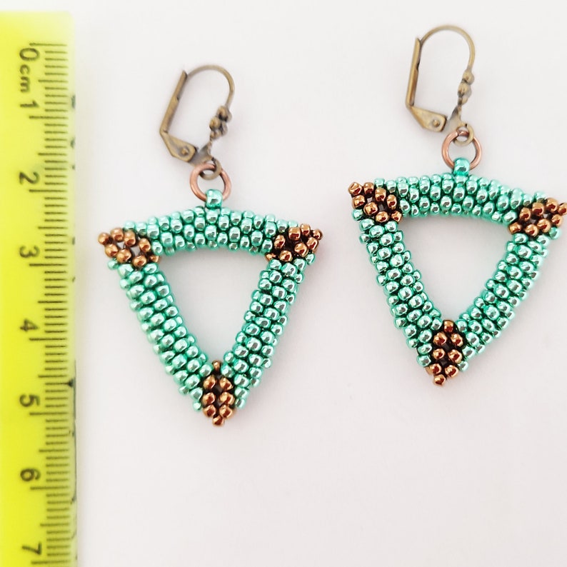 以下が含まれることがあります： 手作りのイヤリングで、三角形のデザインが特徴です。イヤリングは、小さな淡い緑色のビーズで作られており、角にはブロンズ色のビーズが付いています。ブロンズ色のレバーバック留め具が付いています。