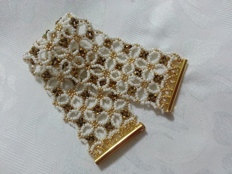 Puede incluir: Un brazalete de cuentas doradas y blancas con un delicado patr&oacute;n floral. El brazalete tiene cierres de metal dorado en cada extremo.