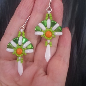 Op de afbeelding: Handgemaakte oorbellen met een waaiervormig ontwerp, bestaande uit groene en witte kralen. Het midden van elke oorbel heeft een oranje kralencluster en een witte druppel hangt eronder. De oorbellen worden in een hand gehouden.