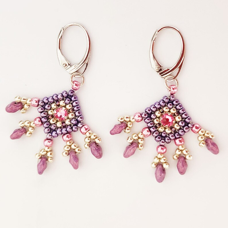 K&ouml;nnte beinhalten: Ein Paar Ohrringe mit geometrischem Design. Die Ohrringe sind aus lila, rosa und goldenen Perlen gefertigt und haben einen rosa Kristall in der Mitte. Die Ohrringe sind an silbernen Haken befestigt.