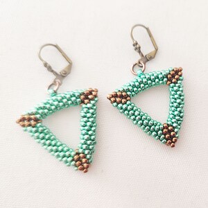 以下が含まれることがあります： 手作りのイヤリングのペアで、三角形のデザインが特徴です。イヤリングは、小さな明るいティールビーズで作られ、角にはブロンズ色のビーズが付いています。イヤリングには、ブロンズ色のレバーバッククロージャーが付いています。