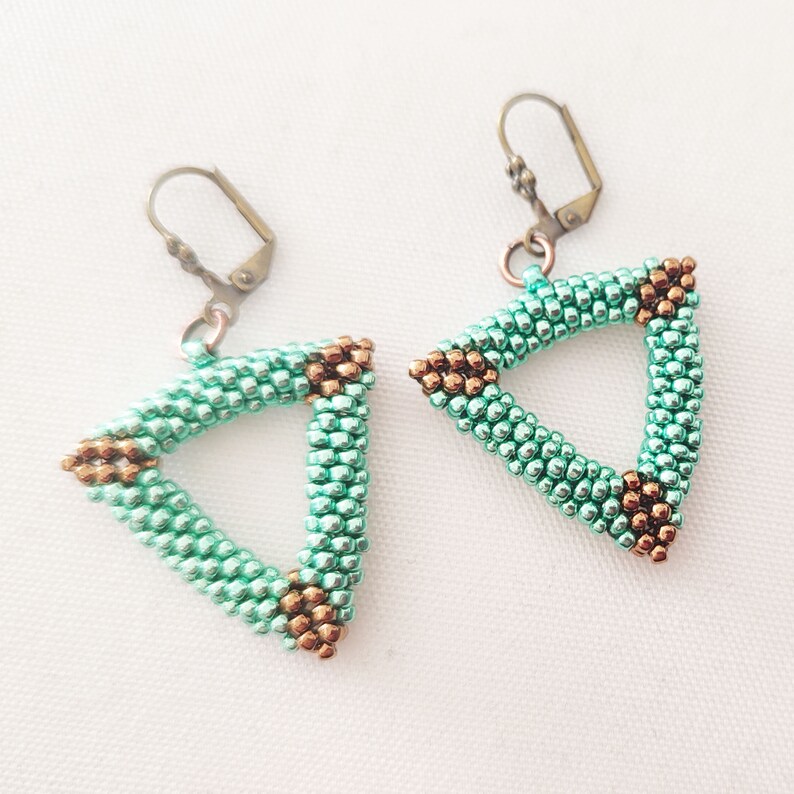 以下が含まれることがあります： 手作りのイヤリングのペアで、三角形のデザインが特徴です。イヤリングは、小さな明るいティールビーズで作られ、角にはブロンズ色のビーズが付いています。イヤリングには、ブロンズ色のレバーバッククロージャーが付いています。