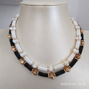 Puede incluir: Un collar de doble cadena con cuentas negras, blancas y doradas. El collar presenta pequeñas cuentas en forma de corazón y está superpuesto con una cadena de cuentas negras y blancas.