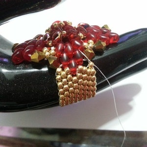 May include: A red and gold beaded jewelry piece with a square base and a pointed top. The piece is made with small, round beads and is being held by a hand.