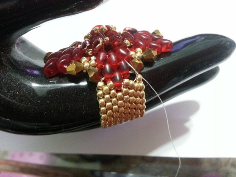 May include: A red and gold beaded jewelry piece with a square base and a pointed top. The piece is made with small, round beads and is being held by a hand.