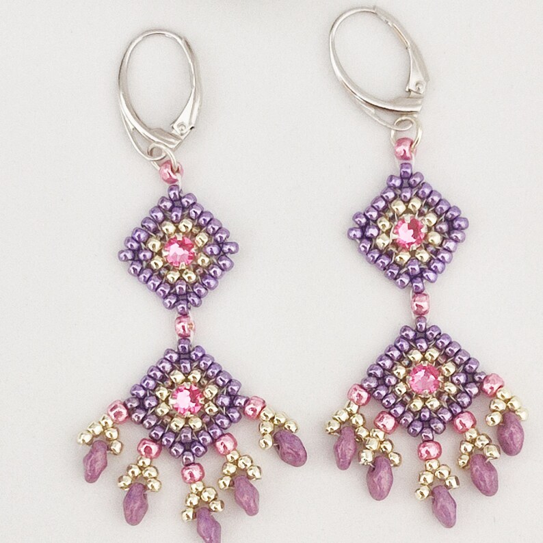 K&ouml;nnte beinhalten: Lila und rosa Perlenohrringe mit quadratischem Design und einem Kristall in der Mitte. Die Ohrringe sind baumelnd und haben einen silbernen Haken.