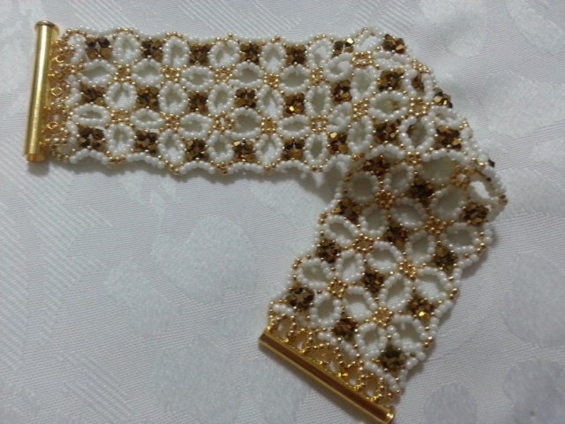 Puede incluir: Un brazalete de cuentas blancas y doradas con un dise&ntilde;o floral. El brazalete est&aacute; hecho con cuentas peque&ntilde;as y redondas y tiene un cierre dorado.