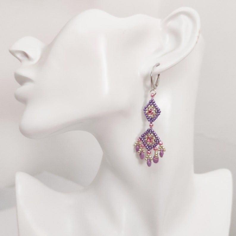 K&ouml;nnte beinhalten: Lila und rosa Perlen-Ohrringe mit einem silbernen Haken. Die Ohrringe haben ein geometrisches Design mit einer kleinen rosa Perle in der Mitte jedes Quadrats.