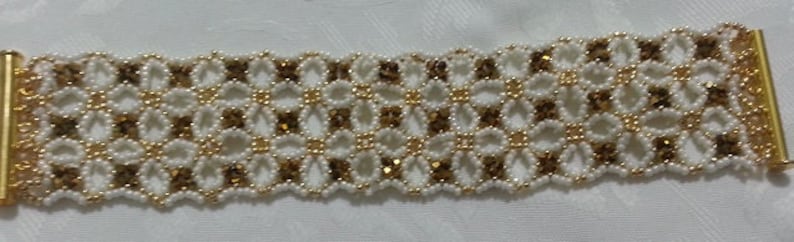 Puede incluir: Un brazalete de cuentas doradas y blancas con un patr&oacute;n geom&eacute;trico. El brazalete est&aacute; hecho con cuentas peque&ntilde;as y redondas y tiene un cierre de broche.