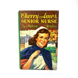 May include: Vintage book cover for "Cherry Ames, Senior Nurse" by Helen Wells. The cover features a smiling nurse in a white cap and navy blue cape. The title is in yellow script on a brown background with green edges.