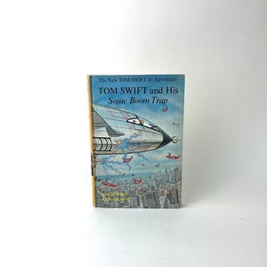 May include: A vintage paperback book cover featuring a blue and white illustration of a futuristic airplane flying over a city skyline. The book title is "TOM SWIFT and His Sonic Boom Trap" and the author is "VICTOR APPLETON II".
