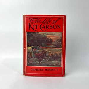 May include: A vintage book titled "The Life of Kit Carson" with a red cover. The cover features a color illustration of a covered wagon pulled by horses, with text in black. The author's name, Charles Burdett, is also printed on the cover.