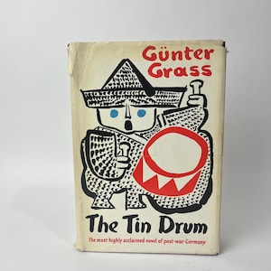 May include: A book titled The Tin Drum by Günter Grass with a cream-colored cover. The front features a black, white, red, and blue graphic illustration of a figure wearing a hat and holding a drum. The text reads Günter Grass, The Tin Drum, and The most highly acclaimed novel of post-war Germany.