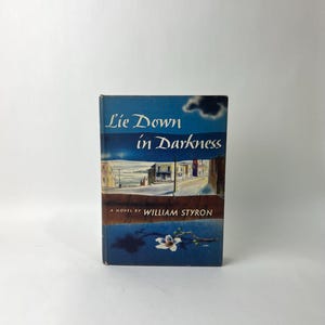 May include: A vintage hardcover book titled "Lie Down in Darkness" by William Styron. The cover features a blue sky with clouds, a street scene, and a white flower. The book is set against a white background.