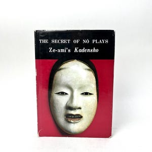 May include: A book titled "THE SECRET OF NŌ PLAYS Ze-ami's Kadensho" with a red cover and a black top. The cover features a white Noh mask with black eyes and a slightly open mouth.