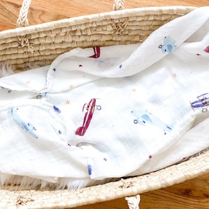 May include: White baby blanket with a pattern of airplanes in blue and burgundy, along with small orange and blue stars. The blanket is folded inside a woven basket with a white fluffy interior.