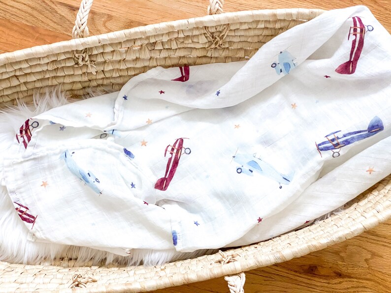 May include: White baby blanket with a pattern of airplanes in blue and burgundy, along with small orange and blue stars. The blanket is folded inside a woven basket with a white fluffy interior.