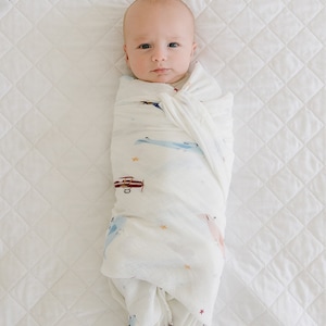 May include: A baby swaddled in a white muslin blanket with a pattern of airplanes, stars, and clouds. The baby is lying on a white quilted surface.