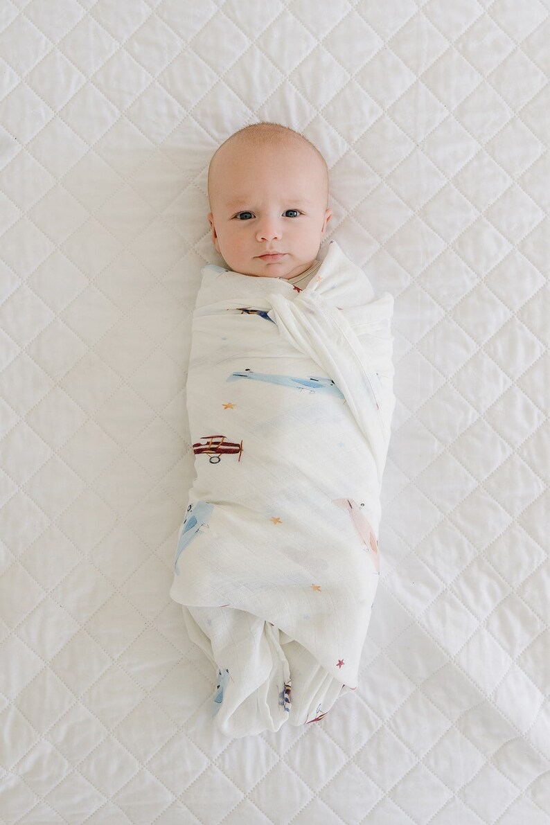 May include: A baby swaddled in a white muslin blanket with a pattern of airplanes, stars, and clouds. The baby is lying on a white quilted surface.