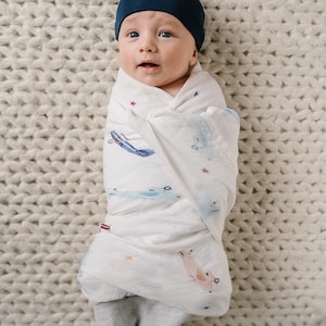 May include: A newborn baby wrapped in a white swaddle blanket with blue and pink airplane illustrations and star accents. The baby wears a navy blue knotted cap and lies on a cream-colored knitted blanket.