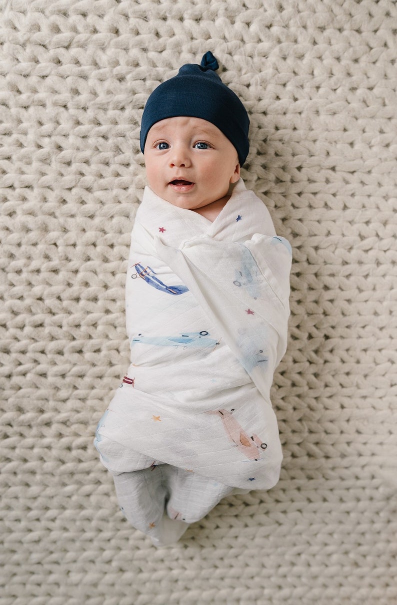 May include: A newborn baby wrapped in a white swaddle blanket with blue and pink airplane illustrations and star accents. The baby wears a navy blue knotted cap and lies on a cream-colored knitted blanket.