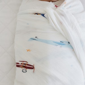 May include: A baby wrapped in a white muslin swaddle blanket. The blanket features a watercolor design of airplanes in shades of blue and maroon, with small orange stars scattered across the fabric. The baby's face is partially visible.