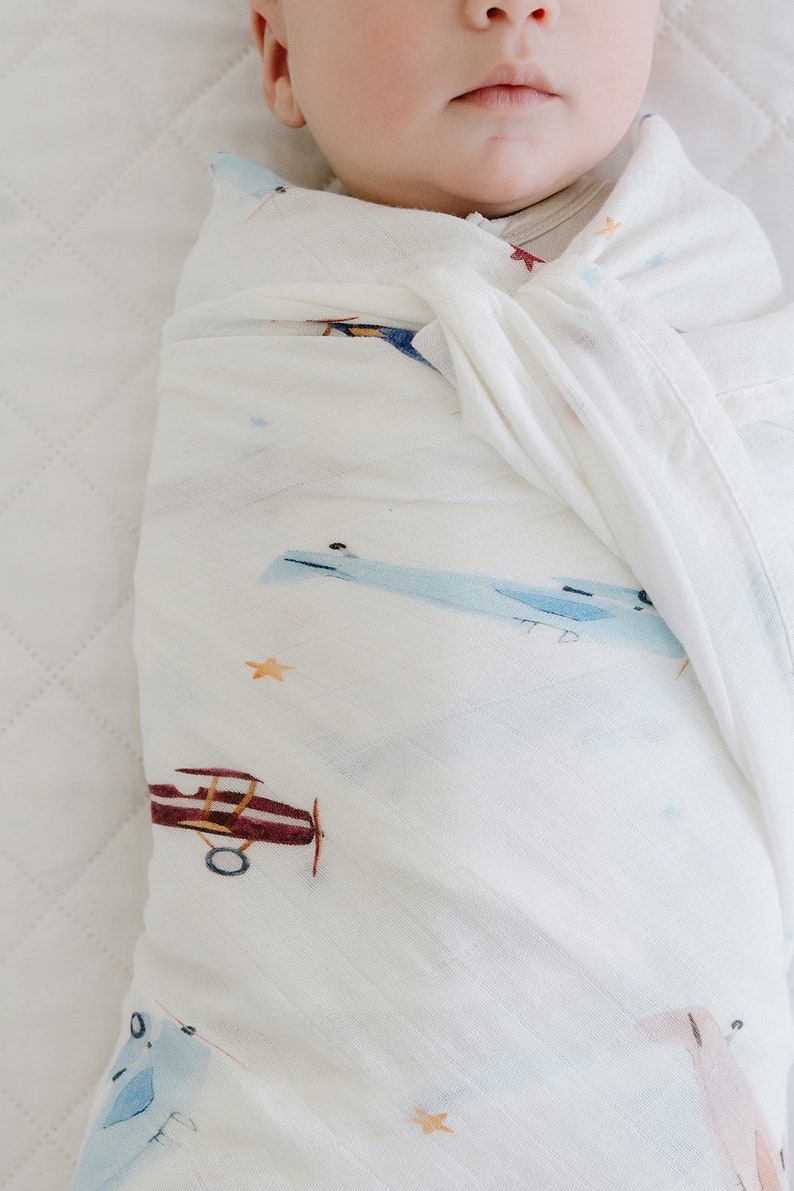 May include: A baby wrapped in a white muslin swaddle blanket. The blanket features a watercolor design of airplanes in shades of blue and maroon, with small orange stars scattered across the fabric. The baby's face is partially visible.