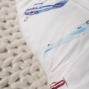 May include: White muslin baby blanket with a watercolor airplane pattern in shades of blue, red, and orange. The blanket is soft and lightweight, perfect for swaddling or as a stroller cover. The background is a chunky knit texture.
