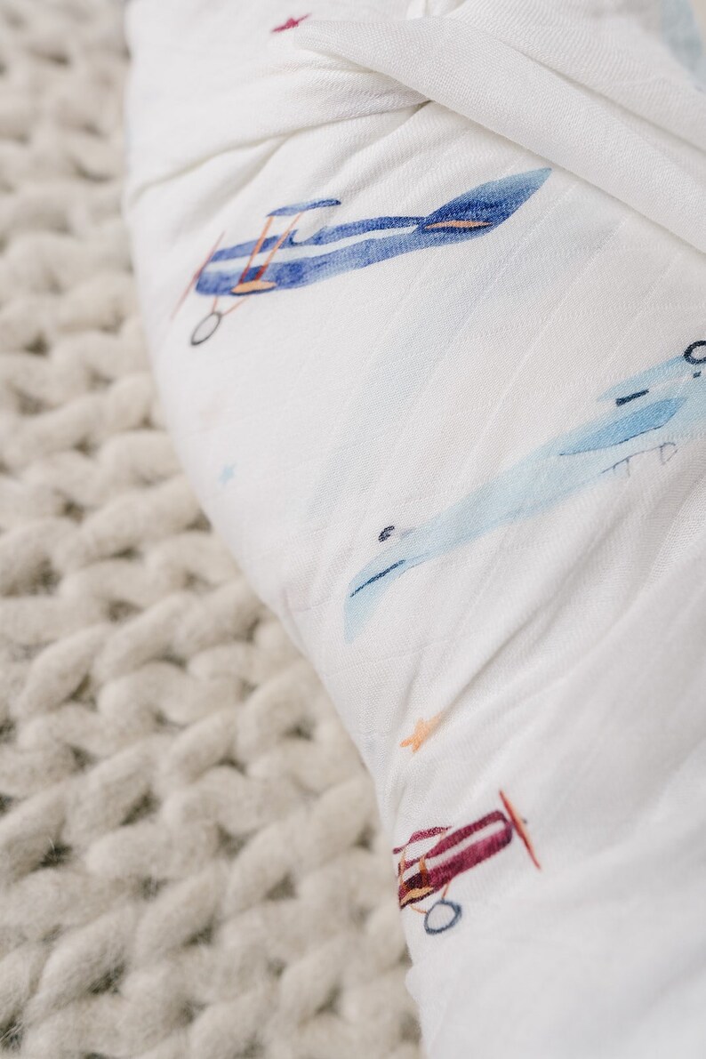 May include: White muslin baby blanket with a watercolor airplane pattern in shades of blue, red, and orange. The blanket is soft and lightweight, perfect for swaddling or as a stroller cover. The background is a chunky knit texture.