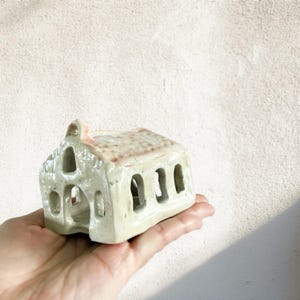 May include: A small, ceramic house-shaped candle holder. The off-white structure has a textured roof and arched openings. The miniature house is held in a hand, showcasing its size and design. The candle holder is a decorative item.