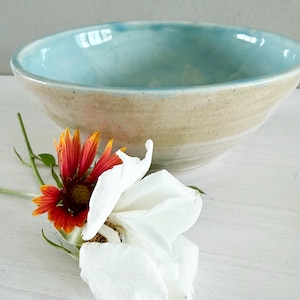 May include: A handmade ceramic bowl with a light blue interior and a beige exterior. The bowl is round and has a wide rim. A red and yellow flower and a white flower are in the foreground.