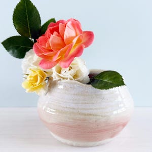 May include: A small, round ceramic vase with a pink band at the base, holding a bouquet of roses. The roses are a mix of colors, including pink, yellow, and white, with green leaves. The vase has a textured, off-white finish.