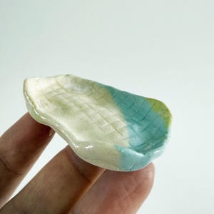 May include: A small, irregular, hand-formed ceramic dish with a glossy finish. The surface features a textured, woven-like grid pattern. The color palette transitions from a creamy off-white to a vibrant turquoise and a soft olive green.