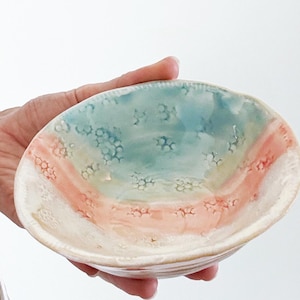 May include: A handmade ceramic bowl with a unique design. The bowl features a color palette of light blue, peach, and white. The interior has a textured surface with a speckled pattern. The bowl is held in a person's hand.