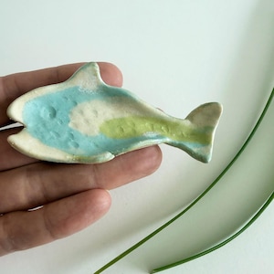 May include: A small, hand-formed ceramic dish shaped like a fish. The surface features a glazed finish with blended patches of light blue, cream, and pale green. The texture is slightly uneven with subtle indentations along the body and tail.