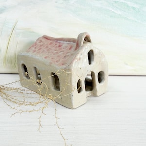 May include: A small, ceramic house with a pink roof and multiple window cutouts. The house is off-white with a textured surface and a handle on top. The background is a soft, blurred painting with green and white hues.