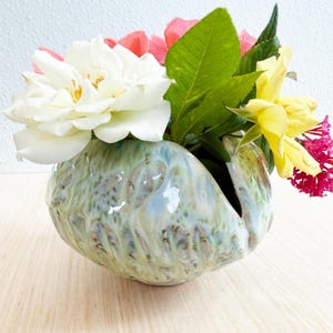 May include: A round, organic-shaped ceramic vase with a glossy, mottled glaze in shades of pale green, blue, and cream. The vase features a deep, vertical slit opening and is filled with a bouquet of white, yellow, pink, and red flowers.