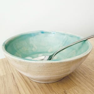 May include: A handmade ceramic bowl with a light blue interior and a beige exterior. A silver spoon rests inside the bowl. The bowl is on a wooden surface. The bowl is approximately 6 inches in diameter.
