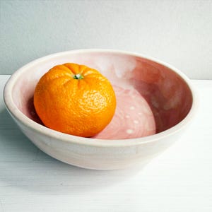 May include: A ceramic bowl with a light pink interior and a white exterior holds a bright orange fruit. The bowl has a smooth, rounded shape and sits on a white surface. The fruit is a vibrant orange color.