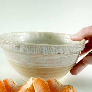 May include: A handmade ceramic bowl with a textured rim and a light beige and white glaze. The bowl has a wide opening and a rounded shape. The bowl is partially filled with orange segments. The bowl is being held by a hand.