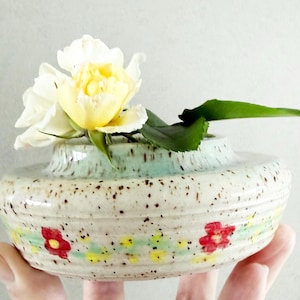 May include: A small, speckled ceramic vase with a light blue rim and floral design. The vase holds a white and yellow rose with green leaves. The vase is held in a person's hand.