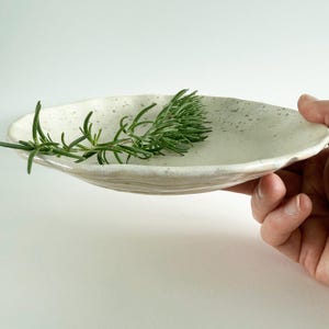 May include: A handmade, off-white ceramic serving dish with a speckled surface and a wavy rim. A sprig of fresh rosemary rests on the dish. The dish is held by a hand against a white background.