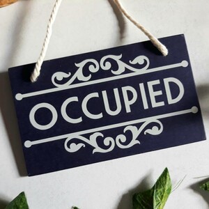 Puede incluir: Un letrero rectangular azul marino con la palabra "OCCUPIED" en blanco, enmarcado por adornos y l&iacute;neas decorativas. El letrero cuelga de una cuerda blanca. El letrero est&aacute; rodeado de hojas verdes.