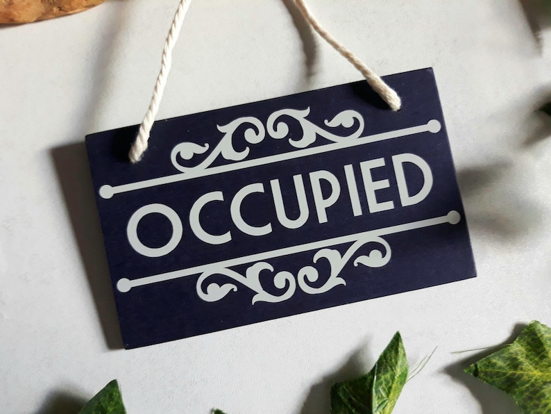 Puede incluir: Un letrero rectangular azul marino con la palabra "OCCUPIED" en blanco, enmarcado por adornos y l&iacute;neas decorativas. El letrero cuelga de una cuerda blanca. El letrero est&aacute; rodeado de hojas verdes.