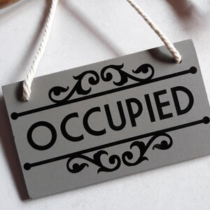 Puede incluir: Un letrero rectangular gris con la palabra "OCCUPIED" en letras negras en negrita. El letrero tiene adornos decorativos negros en la parte superior e inferior y una cuerda blanca para colgar.