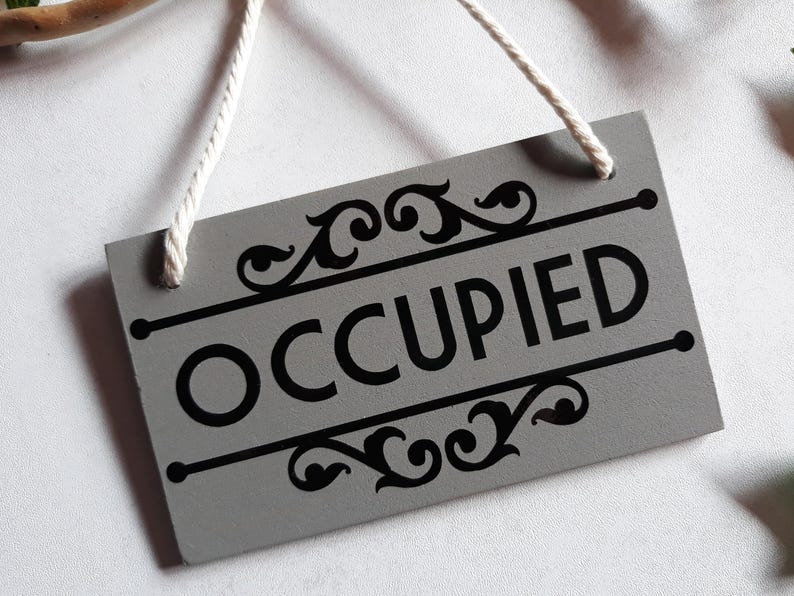 Puede incluir: Un letrero rectangular gris con la palabra "OCCUPIED" en letras negras en negrita. El letrero tiene adornos decorativos negros en la parte superior e inferior y una cuerda blanca para colgar.